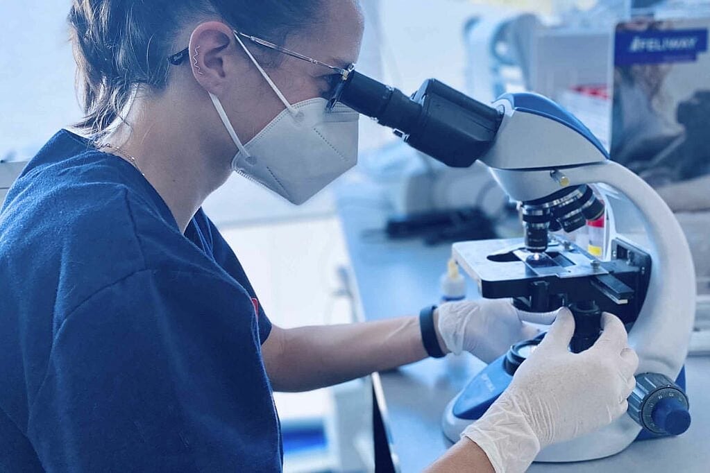 Una persona con mascarilla y guantes mirando a través de un microscopio.
